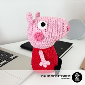 Puede incluir: Juguete de cerdo de crochet rosa con ojos negros y un vestido rojo. El juguete está sentado sobre una superficie blanca con un portátil y cuadernos en el fondo.