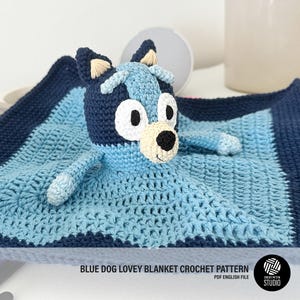 Puede incluir: Manta de crochet azul para bebé con un perro de crochet azul y blanco adjunto. La manta tiene un borde azul y un fondo blanco. El perro tiene una cara blanca con ojos negros y una nariz negra. El texto "BLUE DOG LOVEY BLANKET CROCHET PATTERN" está impreso debajo de la imagen.