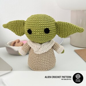 K&ouml;nnte beinhalten: Geh&auml;kelte Figur einer Figur mit gro&szlig;en Ohren, schwarzen Augen und einem gr&uuml;nen Kopf. Die Figur tr&auml;gt eine beige Robe mit cremefarbenem Kragen. Der Text "ALIEN CROCHET PATTERN PDF ENGLISH FILE" befindet sich unten.