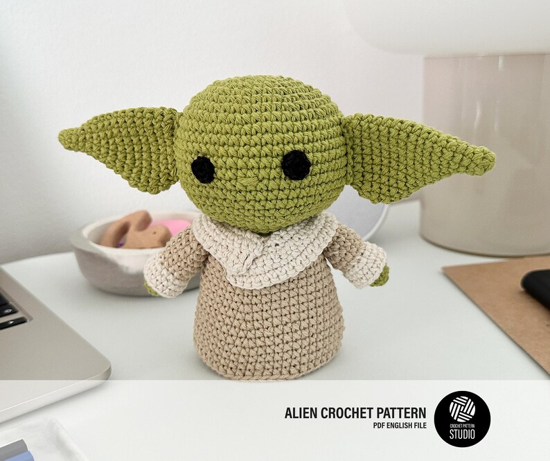 K&ouml;nnte beinhalten: Geh&auml;kelte Figur einer Figur mit gro&szlig;en Ohren, schwarzen Augen und einem gr&uuml;nen Kopf. Die Figur tr&auml;gt eine beige Robe mit cremefarbenem Kragen. Der Text "ALIEN CROCHET PATTERN PDF ENGLISH FILE" befindet sich unten.