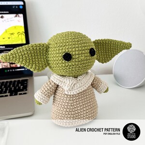 K&ouml;nnte beinhalten: Geh&auml;kelte gr&uuml;ne Alien-Puppe mit gro&szlig;en Ohren, schwarzen Augen und einem beigefarbenen Gewand. Die Puppe sitzt auf einer wei&szlig;en Oberfl&auml;che. Der Text "ALIEN CROCHET PATTERN PDF ENGLISH FILE" ist sichtbar.