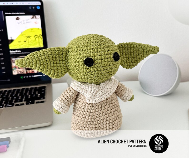 K&ouml;nnte beinhalten: Geh&auml;kelte gr&uuml;ne Alien-Puppe mit gro&szlig;en Ohren, schwarzen Augen und einem beigefarbenen Gewand. Die Puppe sitzt auf einer wei&szlig;en Oberfl&auml;che. Der Text "ALIEN CROCHET PATTERN PDF ENGLISH FILE" ist sichtbar.