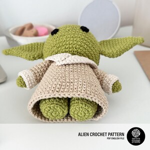 K&ouml;nnte beinhalten: Handgefertigte H&auml;kelpuppe eines Aliens in Gr&uuml;n und Beige. Die Puppe hat gro&szlig;e Ohren, einen beigen Umhang und kleine F&uuml;&szlig;e. Der Text "ALIEN CROCHET PATTERN" ist unten im Bild zu sehen.