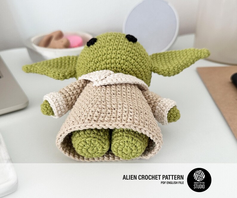 K&ouml;nnte beinhalten: Handgefertigte H&auml;kelpuppe eines Aliens in Gr&uuml;n und Beige. Die Puppe hat gro&szlig;e Ohren, einen beigen Umhang und kleine F&uuml;&szlig;e. Der Text "ALIEN CROCHET PATTERN" ist unten im Bild zu sehen.