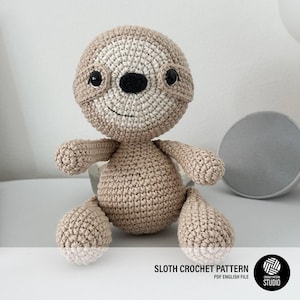 Puede incluir: Un juguete de perezoso tejido a ganchillo con hilo marrón claro y crema, con ojos de plástico negro y nariz negra. El texto SLOTH CROCHET PATTERN PDF ENGLISH FILE está impreso en la parte inferior, junto a un logo circular de Crochet Pattern Studio.