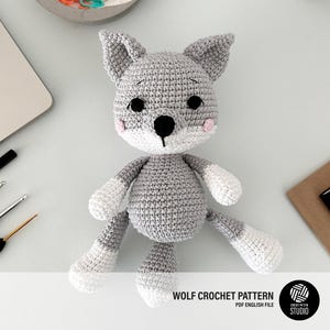 Puede incluir: Un juguete de amigurumi de lobo de crochet gris con detalles blancos en las patas y la cola. El lobo tiene ojos negros y mejillas rosadas. El juguete está sentado sobre una superficie blanca. WOLF CROCHET PATTERN PDF ENGLISH FILE CROCHET PATTERN STUDIO