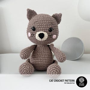 Puede incluir: Un juguete de gato tejido a ganchillo en color marrón con un hocico color crema, nariz negra y grandes ojos negros. El gato tiene mejillas rosadas y orejas puntiagudas. El texto inferior dice: CAT CROCHET PATTERN PDF ENGLISH FILE y un logo.