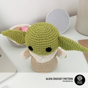 K&ouml;nnte beinhalten: Geh&auml;kelte Alien-Puppe in Gr&uuml;n und Beige, mit gro&szlig;en Ohren und schwarzen Augen. Die Puppe tr&auml;gt ein beiges Outfit und steht auf einer wei&szlig;en Oberfl&auml;che. Der Text lautet "ALIEN CROCHET PATTERN".