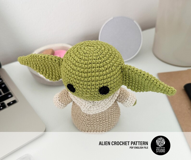 K&ouml;nnte beinhalten: Geh&auml;kelte Alien-Puppe in Gr&uuml;n und Beige, mit gro&szlig;en Ohren und schwarzen Augen. Die Puppe tr&auml;gt ein beiges Outfit und steht auf einer wei&szlig;en Oberfl&auml;che. Der Text lautet "ALIEN CROCHET PATTERN".