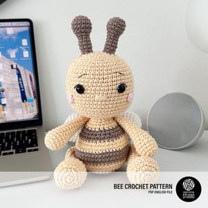Puede incluir: Una muñeca de abeja tejida a ganchillo con cabeza redonda, antenas de color marrón oscuro y cuerpo a rayas en crema y marrón. La muñeca tiene ojos de seguridad negros, mejillas bordadas en rosa y alas blancas. El texto inferior dice BEE CROCHET PATTERN PDF ENGLISH FILE.