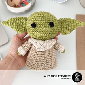 May include: A crocheted alien doll with large green ears, black eyes, and a tan robe. The doll is handmade and has a detailed texture. The image includes text that reads "ALIEN CROCHET PATTERN PDF ENGLISH FILE".