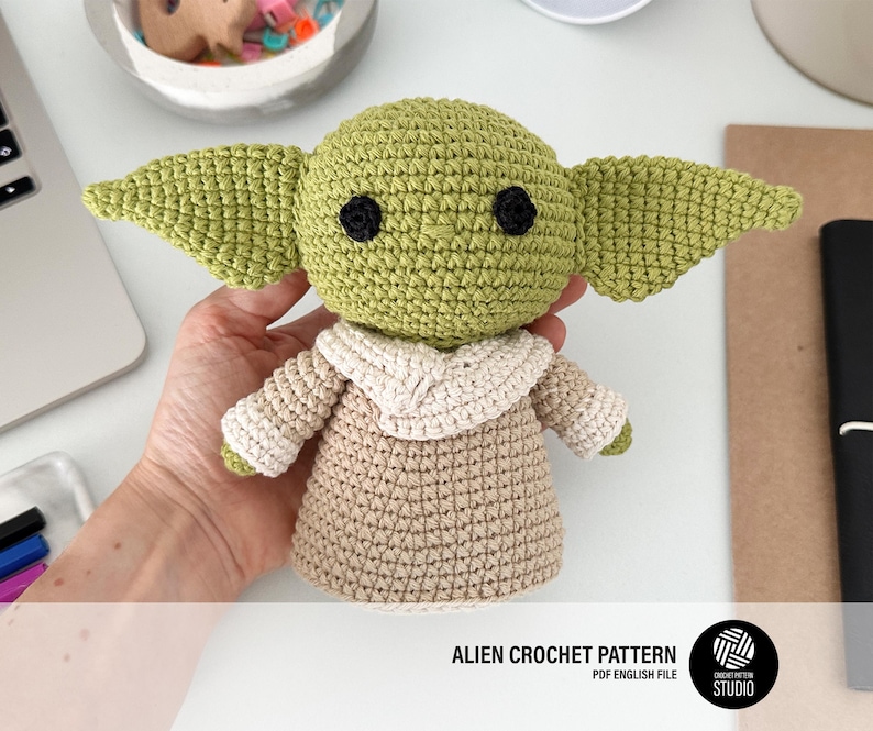 K&ouml;nnte beinhalten: Eine geh&auml;kelte Alien-Puppe mit gro&szlig;en gr&uuml;nen Ohren, schwarzen Augen und einem beigefarbenen Gewand. Die Puppe ist handgefertigt und hat eine detaillierte Textur. Das Bild enth&auml;lt den Text "ALIEN CROCHET PATTERN PDF ENGLISH FILE".