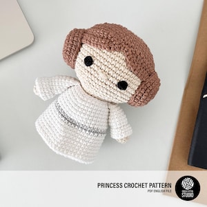 Puede incluir: Muñeca de ganchillo que representa a una princesa, con un vestido blanco con detalles grises y cabello castaño. La muñeca tiene ojos negros y está sobre un fondo claro. Se puede ver el texto "PRINCESS CROCHET PATTERN".