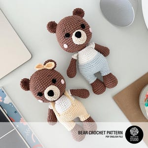 Puede incluir: Dos juguetes de peluche de oso marrón hechos a crochet. Un oso lleva un atuendo azul claro y el otro lleva un atuendo amarillo y blanco. Los osos están sobre una superficie blanca.
