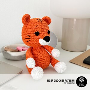 Puede incluir: Un juguete de tigre tejido a ganchillo en naranja y blanco, con detalles negros. El tigre tiene hocico y patas blancas, ojos negros y rayas negras en la cabeza. El texto "TIGER CROCHET PATTERN PDF ENGLISH FILE" es visible.