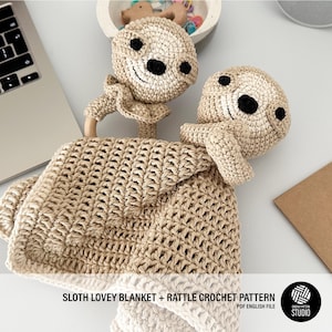 Puede incluir: Un conjunto de manta y sonajero de crochet en forma de perezoso. La manta es beige y presenta un perezoso de crochet con ojos negros. El sonajero es un anillo de madera con una cabeza de perezoso de crochet unida.