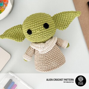 K&ouml;nnte beinhalten: Geh&auml;kelte gr&uuml;ne Alien-Puppe mit gro&szlig;en Ohren, schwarzen Augen und einem beigefarbenen Gewand. Die Puppe befindet sich auf einer hellgrauen Oberfl&auml;che. Der Text "ALIEN CROCHET PATTERN PDF ENGLISH FILE" ist unten im Bild.
