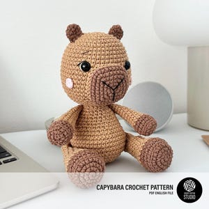 Puede incluir: Un peluche de capibara tejido a ganchillo en tonos beige y marrón con ojos de seguridad negros, nariz bordada y pequeños detalles circulares rosas en las mejillas. El texto CAPYBARA CROCHET PATTERN PDF ENGLISH FILE aparece abajo a la derecha.