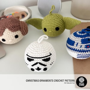 Puede incluir: Adornos navideños de ganchillo con personajes de una popular franquicia de ciencia ficción. Los adornos incluyen un personaje con cabello castaño, un personaje verde con orejas grandes, un adorno blanco con una cara negra y un droide azul y blanco.