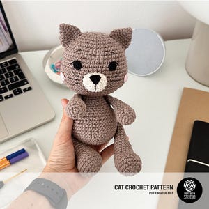 Puede incluir: Un muñeco de gato tejido a ganchillo, en tonos marrones, con ojos negros y hocico blanco. El gato tiene pequeñas mejillas rosas y está siendo sostenido. La imagen también incluye un portátil, un cuaderno y el texto "CAT CROCHET PATTERN PDF ENGLISH FILE".