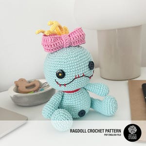 Puede incluir: Un muñeco de trapo de crochet azul con un lazo rosa y pelo amarillo. El muñeco tiene una boca cosida y ojos de botón.