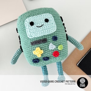 May include: Green crocheted amigurumi toy shaped like a video game console with a screen, buttons, and a joystick. The toy has a smiling face and is made with different colored yarn.