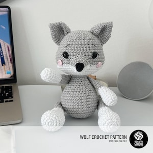May include: A gray and white crocheted wolf plush toy with black eyes, a black nose, and small pink cheeks. The wolf is in a sitting position with one arm raised. The text WOLF CROCHET PATTERN PDF ENGLISH FILE is visible at the bottom right.