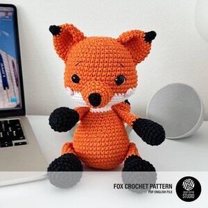 Puede incluir: Un juguete de zorro tejido a ganchillo con hilo naranja brillante, patas negras, puntas de orejas negras y hocico blanco. El zorro tiene grandes ojos de seguridad negros y mejillas bordadas en rosa. El texto dice FOX CROCHET PATTERN PDF ENGLISH FILE.