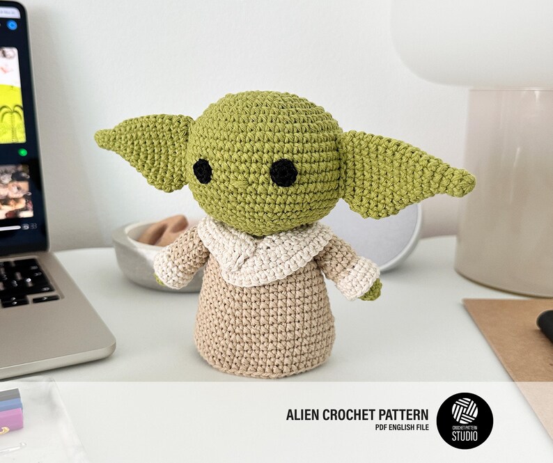 K&ouml;nnte beinhalten: Eine geh&auml;kelte Figur eines Charakters mit gro&szlig;en Ohren, gr&uuml;nem Kopf und schwarzen Augen. Die Figur tr&auml;gt eine beige Robe mit cremefarbenem Kragen. Der Text "ALIEN CROCHET PATTERN PDF ENGLISH FILE" befindet sich am unteren Bildrand.