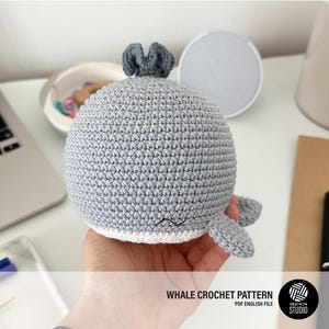 Puede incluir: Un juguete de ballena gris tejido a ganchillo con una pequeña aleta dorsal y aletas caudales. La ballena tiene una barriga blanca y una pequeña boca bordada. El texto "WHALE CROCHET PATTERN" es visible en la imagen, junto con las palabras "PDF ENGLISH FILE".