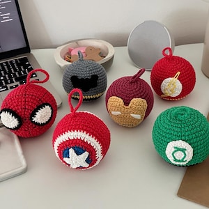 Puede incluir: Una colección de siete adornos de ganchillo, cada uno con un tema de superhéroe diferente. Los adornos son principalmente rojos, verdes, grises y granates, con detalles blancos y amarillos. Cada adorno tiene un lazo para colgar.