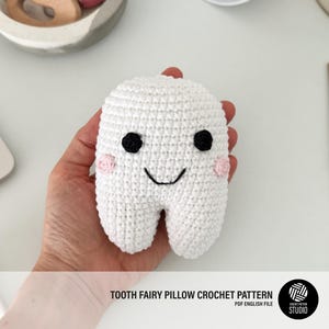 Puede incluir: Una almohada blanca de ganchillo con forma de diente con ojos negros, boca sonriente y mejillas rosas. La almohada está hecha a mano y diseñada para el hada de los dientes. El texto "TOOTH FAIRY PILLOW CROCHET PATTERN" es visible en la parte inferior.