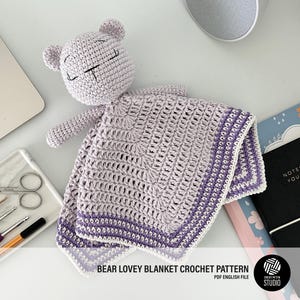 Puede incluir: Una manta de bebé de crochet de color lavanda y blanco con un oso de crochet adjunto. La manta tiene un borde de hilo lavanda y blanco. La manta está sobre una superficie blanca con un cuaderno y otros artículos.