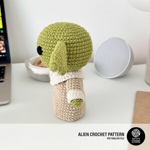 K&ouml;nnte beinhalten: Handgefertigte gr&uuml;ne Alien-H&auml;kelpuppe mit gro&szlig;en Ohren, schwarzen Augen und einem beigen Outfit. Die Puppe sitzt auf einer wei&szlig;en Oberfl&auml;che. Der Text "ALIEN CROCHET PATTERN" befindet sich am unteren Rand des Bildes.