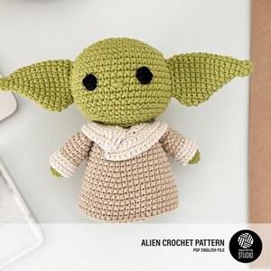 K&ouml;nnte beinhalten: Geh&auml;kelte Alien-Puppe in Gr&uuml;n, Beige und Wei&szlig;. Die Puppe hat gro&szlig;e Ohren und schwarze Augen und tr&auml;gt eine beige Robe mit wei&szlig;em Kragen. Der Text "ALIEN CROCHET PATTERN" ist sichtbar.