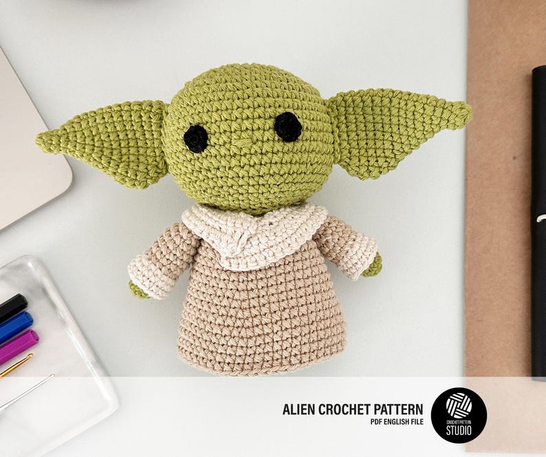 K&ouml;nnte beinhalten: Geh&auml;kelte Alien-Puppe in Gr&uuml;n, Beige und Wei&szlig;. Die Puppe hat gro&szlig;e Ohren und schwarze Augen und tr&auml;gt eine beige Robe mit wei&szlig;em Kragen. Der Text "ALIEN CROCHET PATTERN" ist sichtbar.