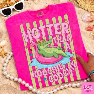 May include: A vibrant pink t-shirt with the phrase "HOTTER THAN A HOOCHIE COOCHIE" in pink and green. The design shows a green alligator in sunglasses on a pink flamingo float. The t-shirt has a crew neck.