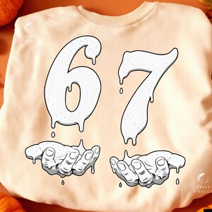 May include: Cream-colored sweatshirt with a graphic design featuring the numbers "67" in a dripping style, above two hands also in a dripping style. The design is in white with black outlines.