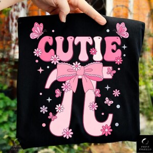 May include: Black t-shirt with a pink design featuring the word "CUTIE" above a pink bow and the mathematical symbol Pi. The design includes pink flowers, butterflies, and stars.