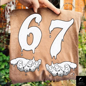 May include: A brown t-shirt featuring the numbers "67" in a dripping white font, with white hands below. The shirt has the text "PRINT INKELLO" in the corners. The design is a graphic illustration.