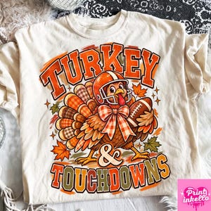 Könnte beinhalten: Ein cremefarbenes T-Shirt mit einer Grafik eines Truthahns, der einen orangefarbenen Football-Helm und eine karierte Fliege trägt und einen Football hält. Der Text Turkey & Touchdowns steht in orangefarbenen und braunen Buchstaben mit Herbstblättern.