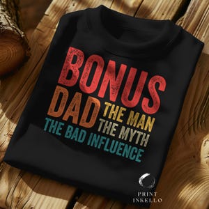 Puede incluir: Sudadera negra de cuello redondo con el texto "BONUS DAD THE MAN THE MYTH THE BAD INFLUENCE" en letras rojas, naranjas, amarillas y turquesas. La sudadera está doblada sobre una superficie de madera.