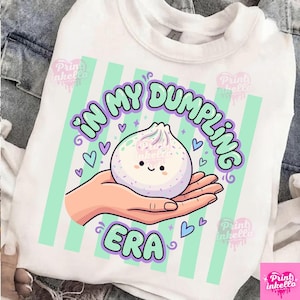 May include: White t-shirt with a graphic of a smiling dumpling held in a hand, surrounded by hearts and the text "IN MY DUMPLING ERA" in a whimsical font. The design features pastel green and purple accents.