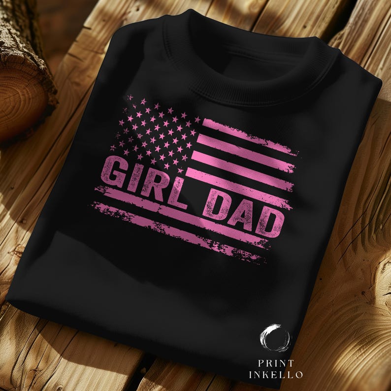 Puede incluir: Sudadera negra con un dise&ntilde;o de bandera estadounidense rosa desgastada. La bandera presenta estrellas y rayas, con las palabras "GIRL DAD" impresas. La sudadera est&aacute; doblada sobre una superficie de madera.