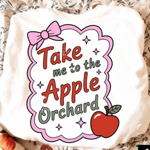 May include: White sweatshirt with a pink bow and a graphic that says "Take me to the Apple Orchard" in red, green, and pink. A red apple with a green leaf is on the bottom right. The design is outlined in pink.