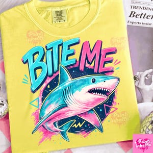 Puede incluir: Camiseta amarilla con el texto "BITE ME" en azul y rosa, con un tibur&oacute;n de dibujos animados con detalles en azul y rosa. La camiseta tiene cuello redondo y una etiqueta Comfort Colors.
