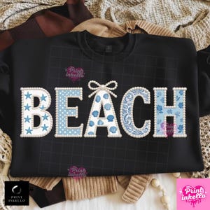 May include: Black sweatshirt with the word "BEACH" in white and blue. The letters are decorated with starfish, polka dots, seashells, and floral patterns. The letters are outlined with white pearls.