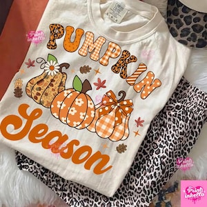 Può includere: Una t-shirt color crema con la scritta PUMPKIN SEASON in arancione. Il design include tre zucche illustrate con vari motivi come quadri, pois e stampa leopardata, arricchite da piccole foglie e stelle nei toni dell'arancione e del marrone.