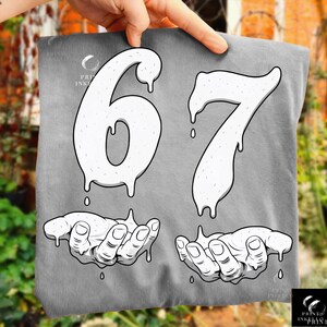 May include: A gray t-shirt featuring the numbers "67" in a dripping, textured white font. Below the numbers are two hands, also in white, with a dripping effect. The shirt has the "Print Ink Print" logo in the upper left and lower right corners.