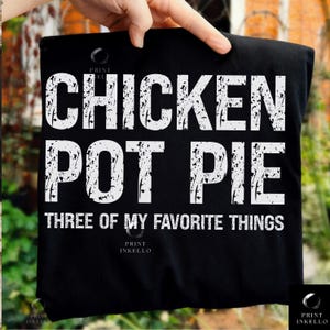 Può includere: T-shirt nero con la scritta bianca "CHICKEN POT PIE THREE OF MY FAVORITE THINGS" in un carattere invecchiato. La maglietta è tenuta in mano, mostrando il design. Il design è semplice e audace.